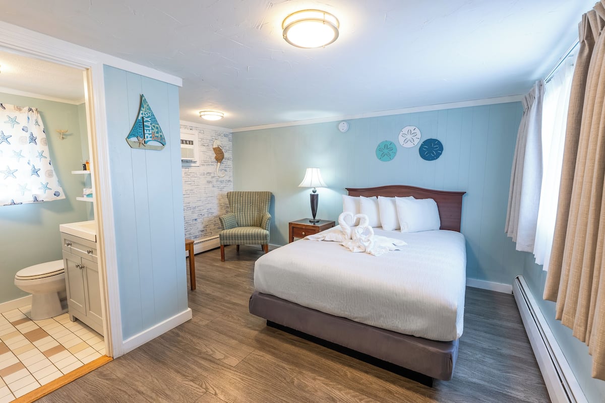 A bedroom with a bed, a chair, a nightstand, a lamp, and a bathroom with a toilet and shower curtain.