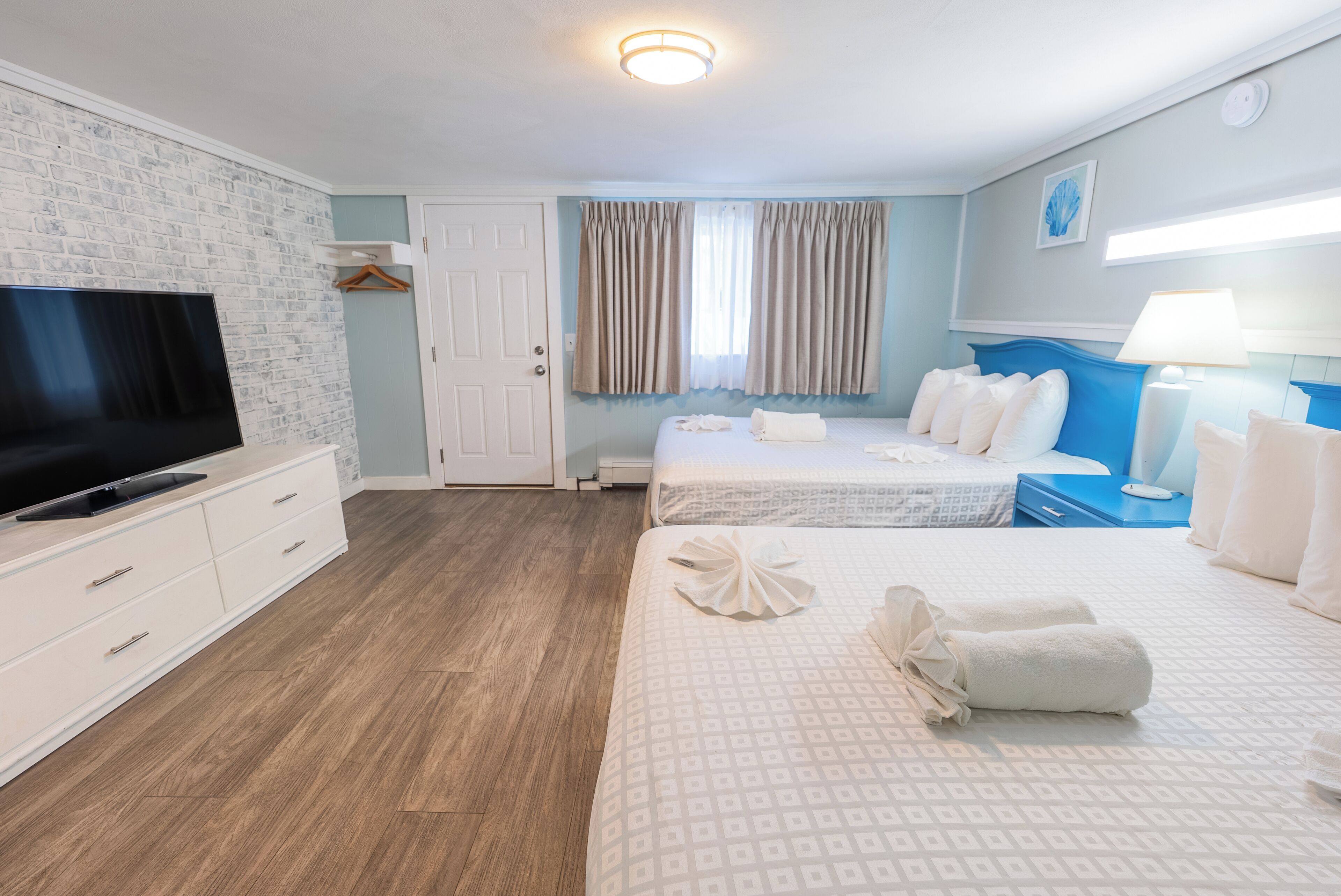 A hotel room with a flat-screen TV, a bed with white linens, a bedside table with a lamp, and a wooden floor.