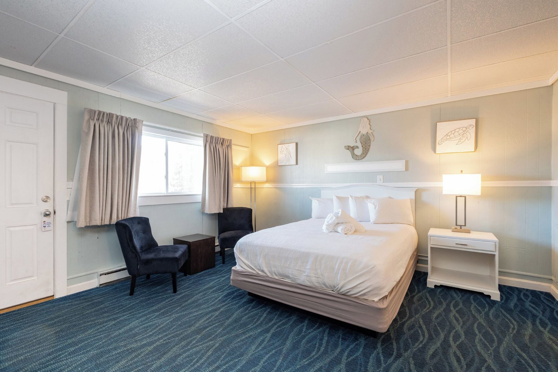 A hotel room with a bed, two chairs, a desk, a lamp, and a window with curtains.