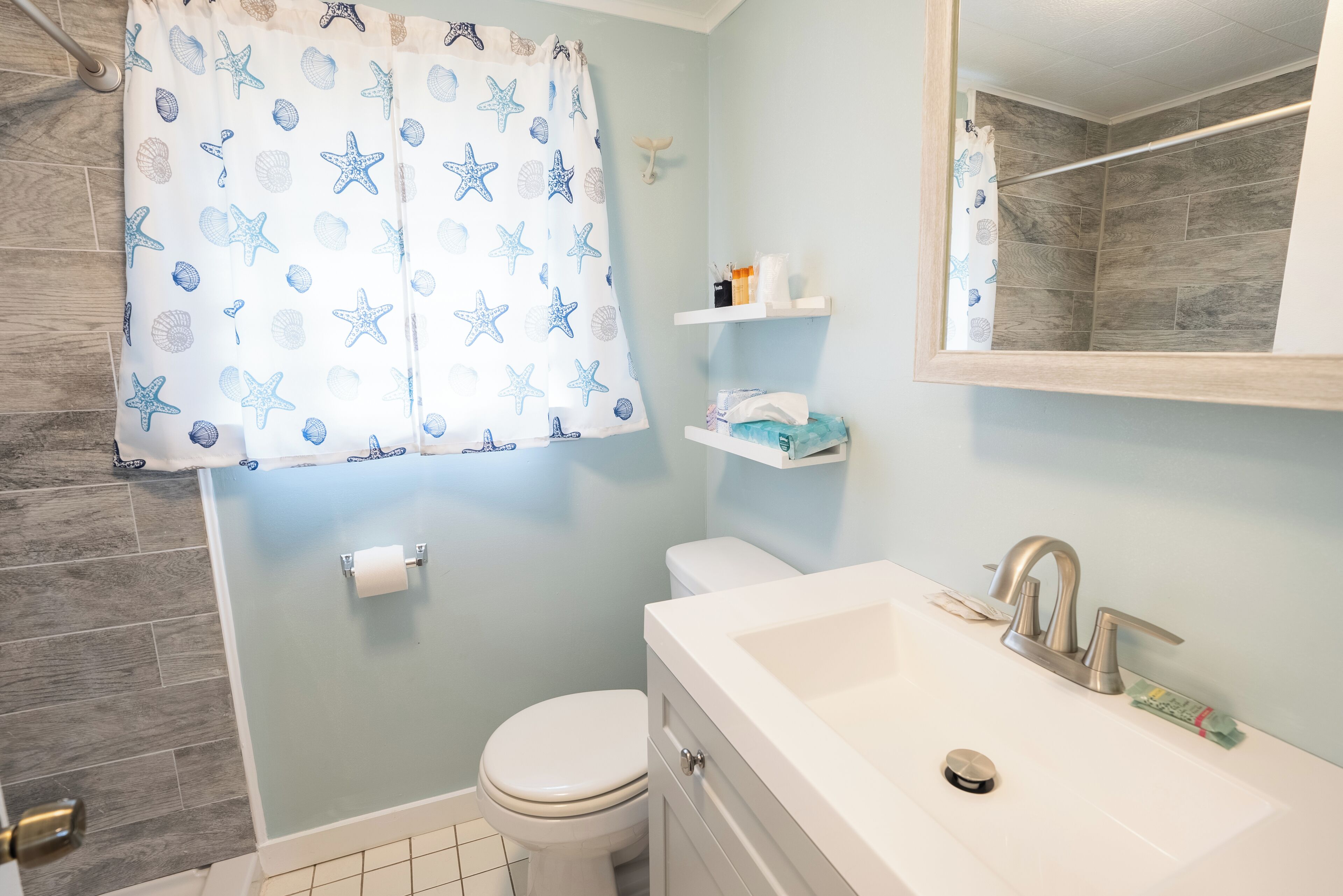A bathroom with a white bathtub, a white toilet, a shower curtain with starfish and shell patterns, and a mirror above the sink.