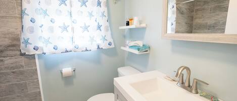 A bathroom with a white bathtub, a white toilet, a shower curtain with starfish and shell patterns, and a mirror above the sink.