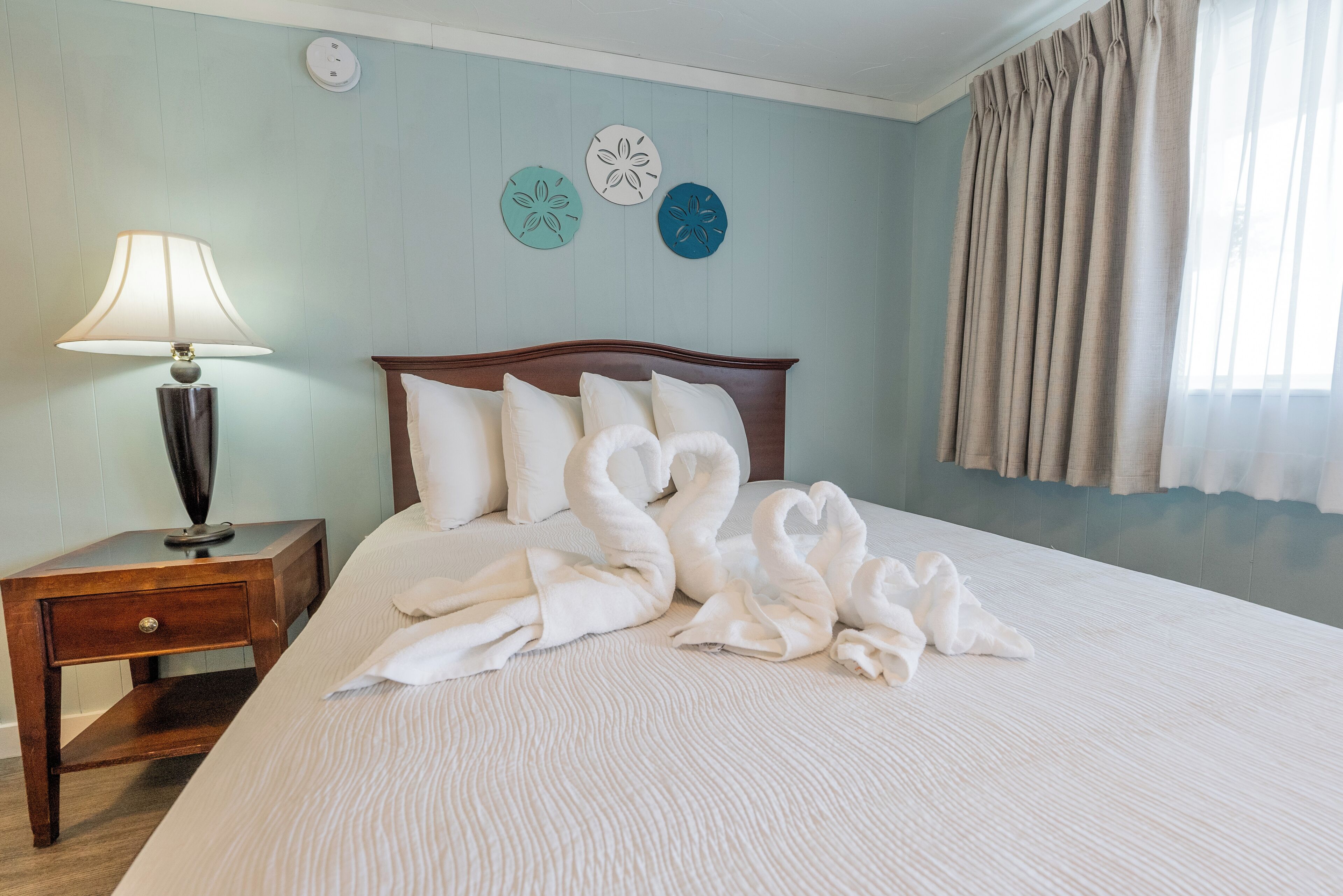 Una cama bien hecha con sábanas blancas y toallas con forma de cisne, una mesita de noche de madera con una lámpara y decoración en la pared.