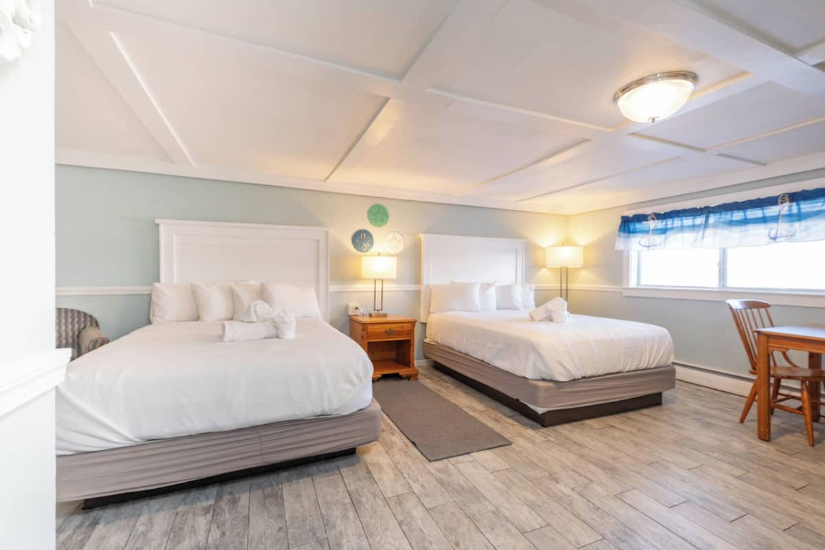 A hotel room with two beds, a wooden floor, a small table, and a window with a blue curtain.