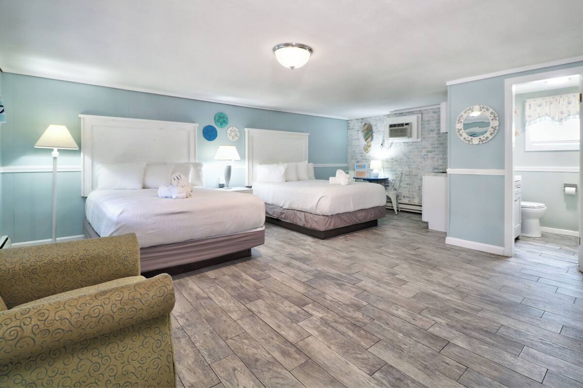 A hotel room with two beds, a sitting area with a patterned chair, a bathroom with a toilet and sink, and a small table with a lamp.