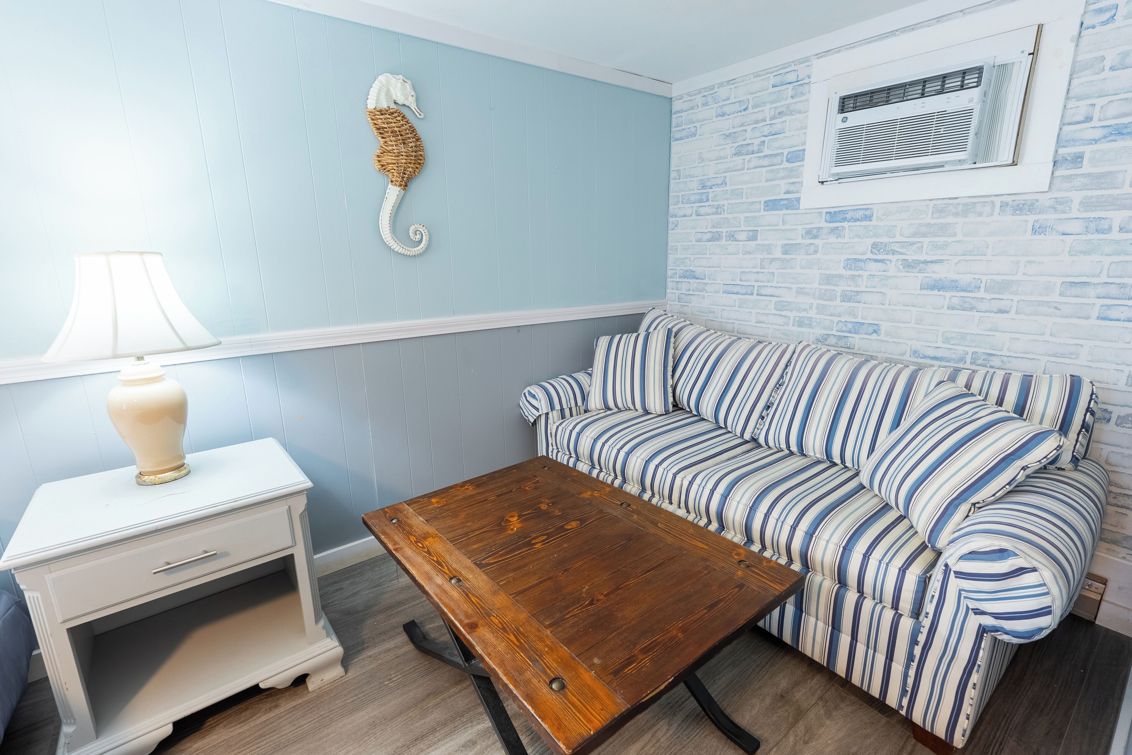 Une pièce avec un canapé à rayures, une table basse en bois, une table de chevet blanche avec une lampe, et un mur décoré d’un motif en briques.
