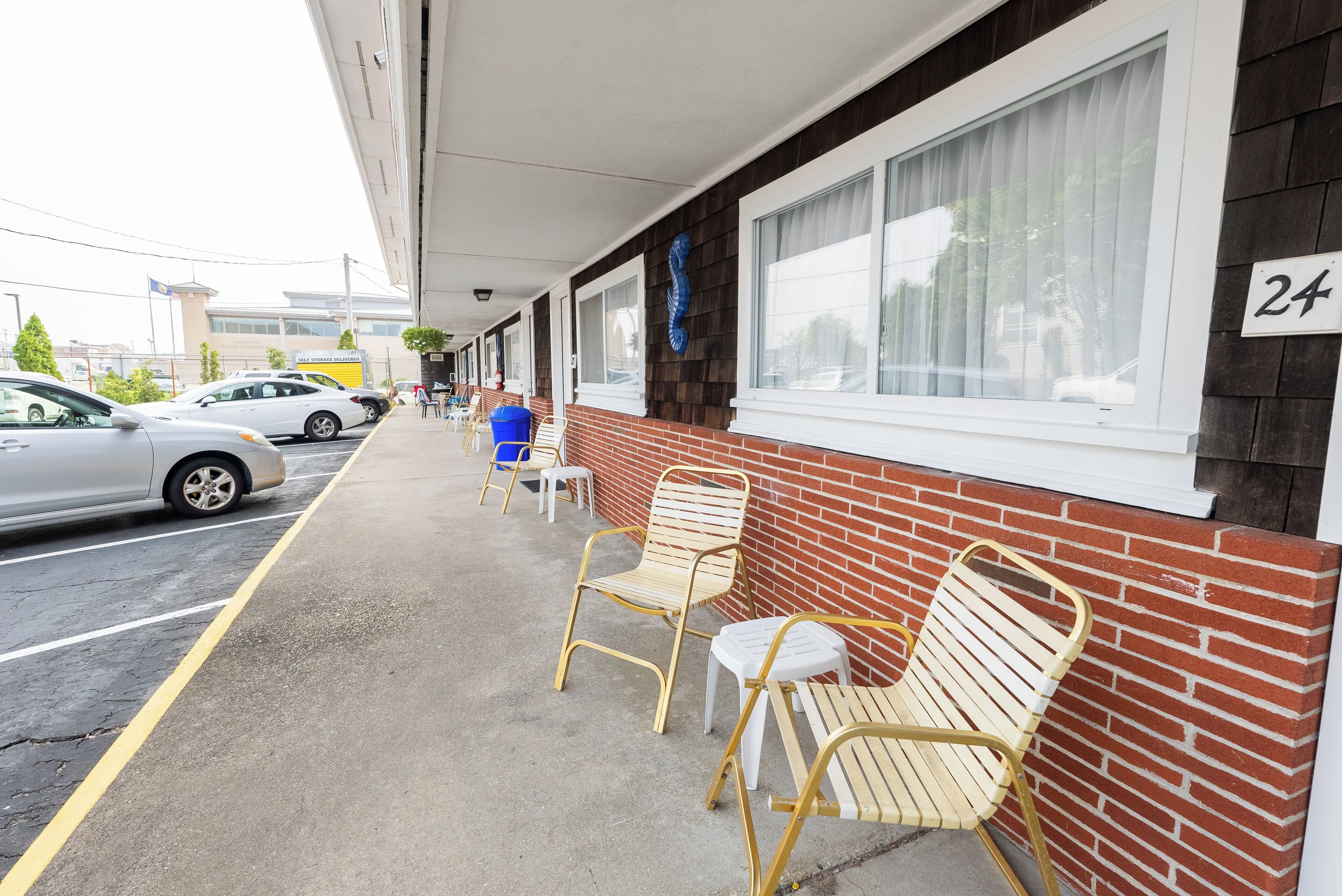 A parking area with cars, a row of outdoor chairs, and a building with a brick facade and large windows.