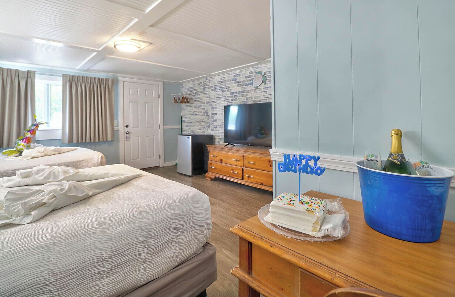 A hotel room with two beds, a television, a minibar, and a birthday cake in a bucket.