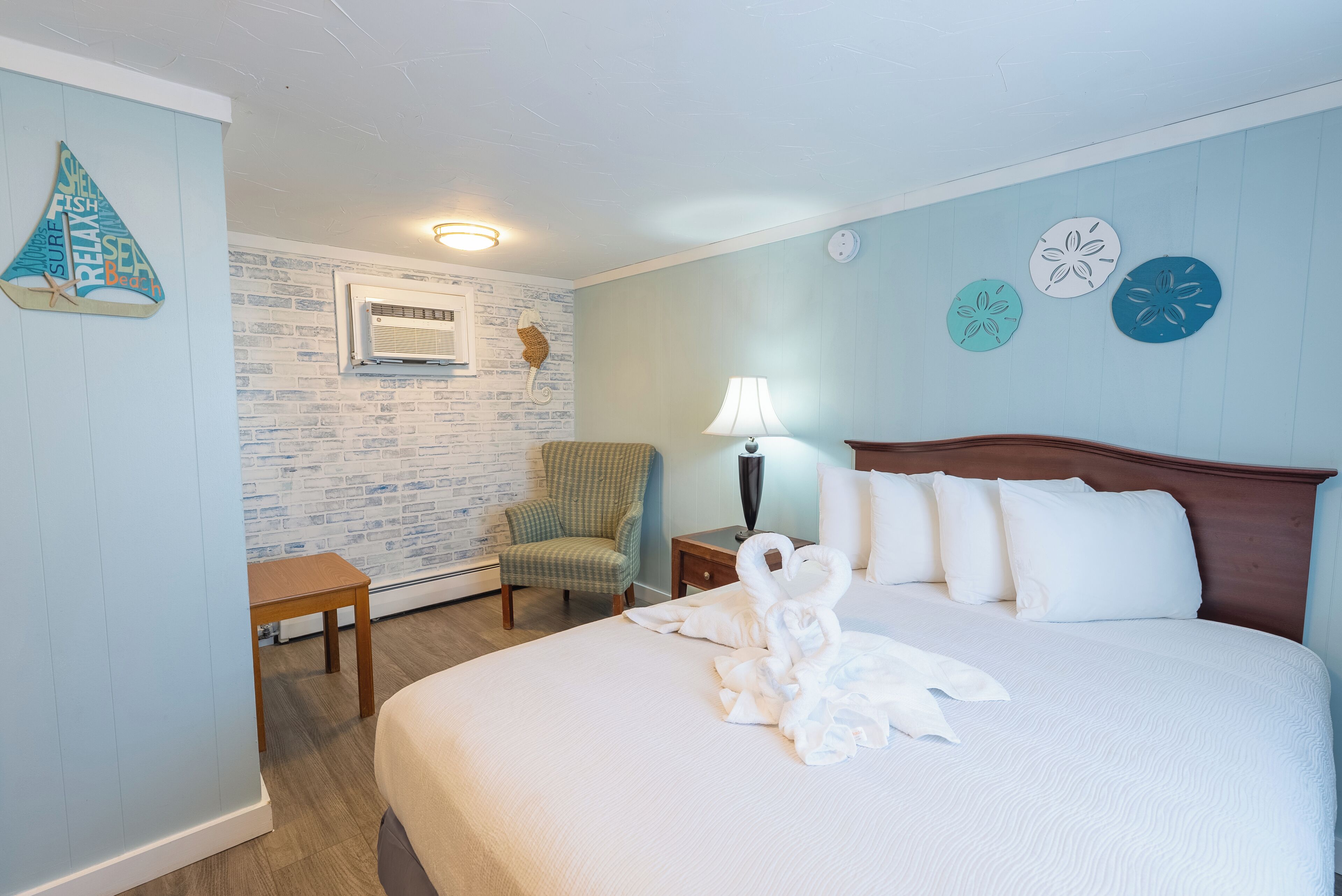 A hotel room with a neatly made bed, a chair, a nightstand with a lamp, and a wall with decorative elements.