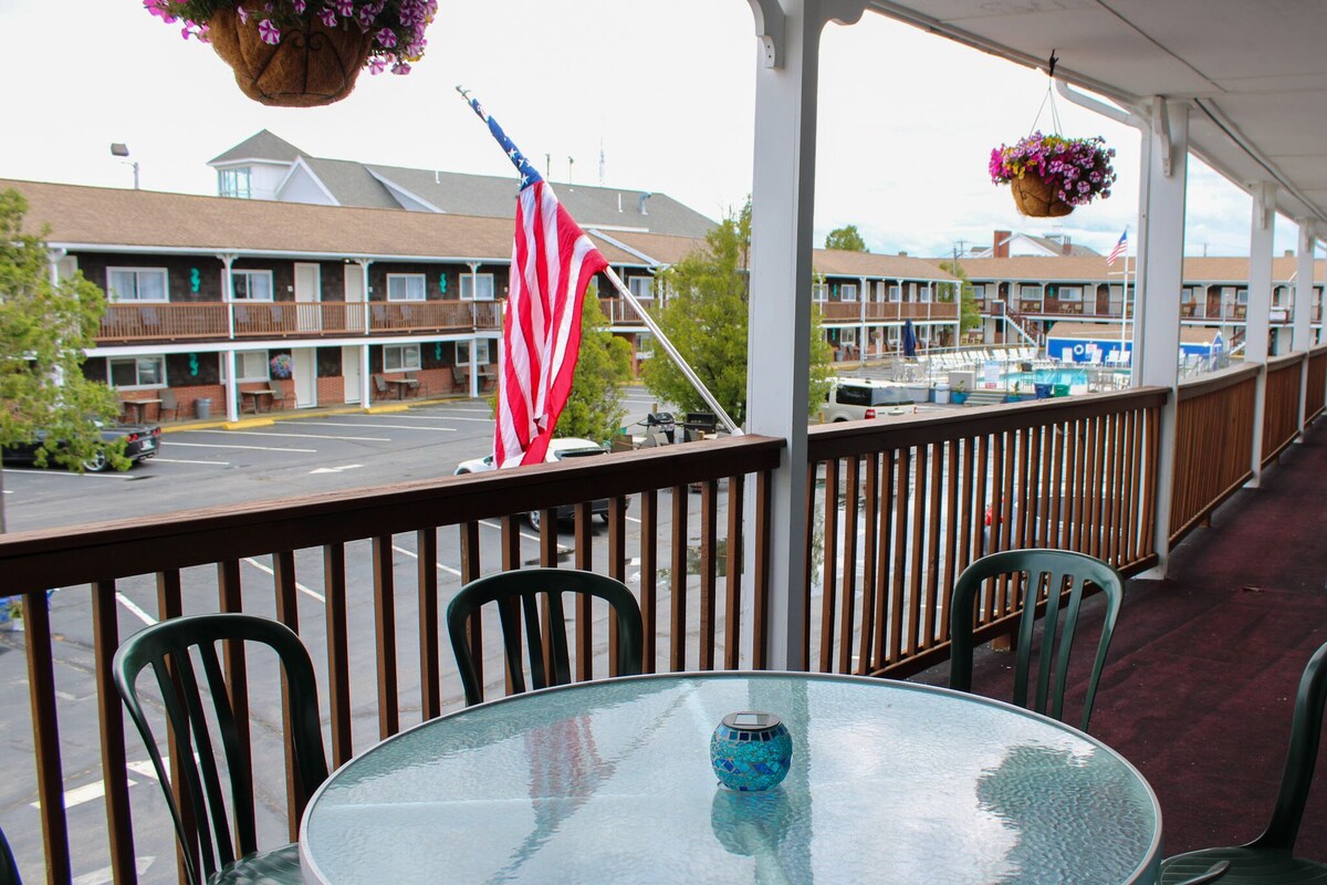 A balcony with a round glass table, chairs, and hanging flower baskets. An American flag is visible on the railing.