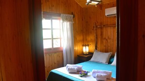 A wooden room with a bed, a nightstand with a lamp, and a window with curtains.