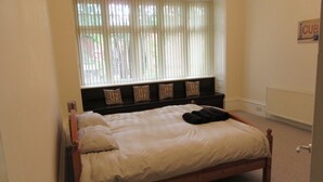 Ein ordentlich bezogenes Bett mit Kissen und Decke, ein Fenster mit Jalousien, ein Heizkörper und ein Schild an der Wand.