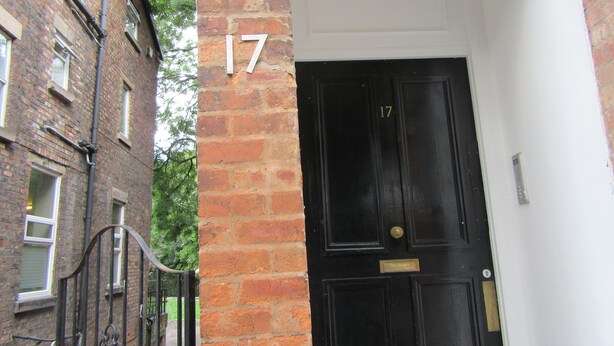 Une porte noire, dotée d’une poignée et d’une plaque en laiton, flanquée d’un pilier en briques portant le numéro 17.