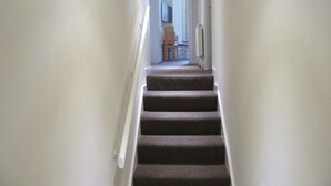 A narrow staircase with carpeted steps leading to a room with a chair and a table.