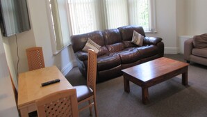 A living room with a brown leather sofa, a wooden coffee table, a wooden chair, and a television mounted on the wall.