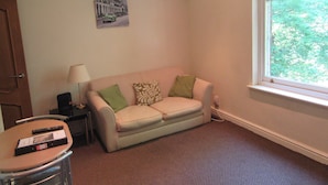 A small, furnished room with a sofa, a lamp, a remote control, and a window with a view of trees. - No 7 (Liverpool)