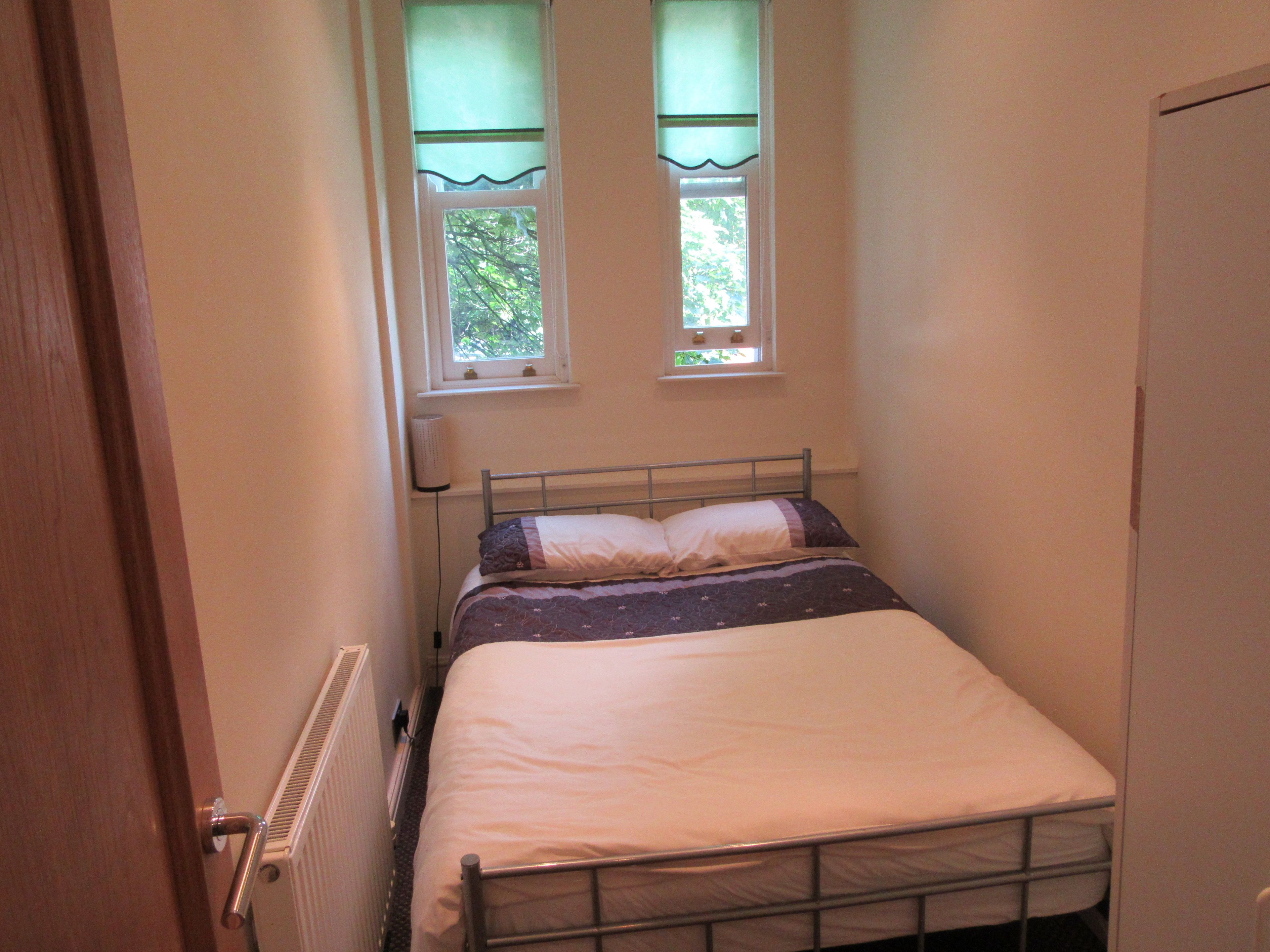 A small, single-bed room with a metal frame, a radiator, and two windows with green curtains.