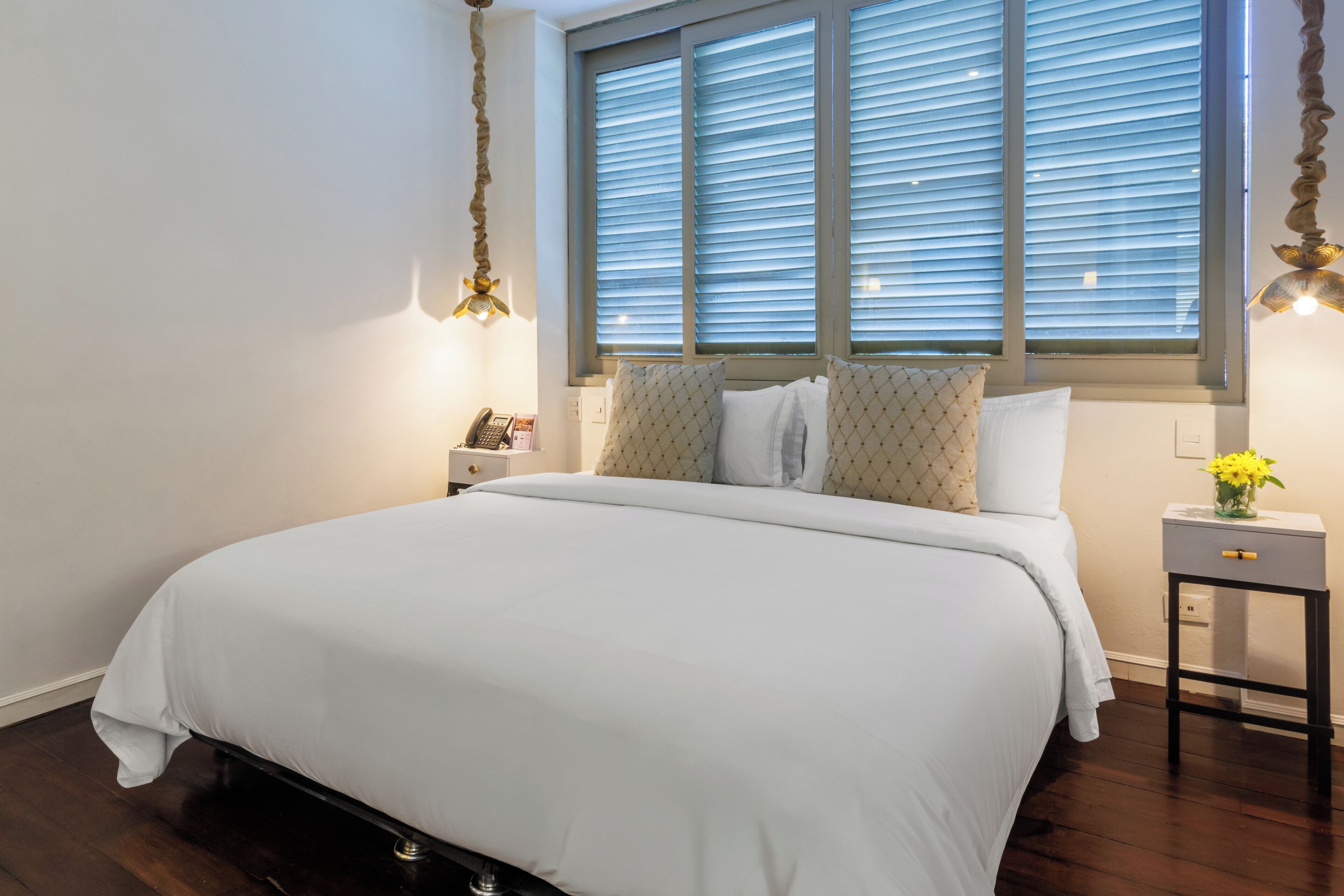 A neatly made bed with a white comforter, a bedside table with a lamp, and a window with blinds.