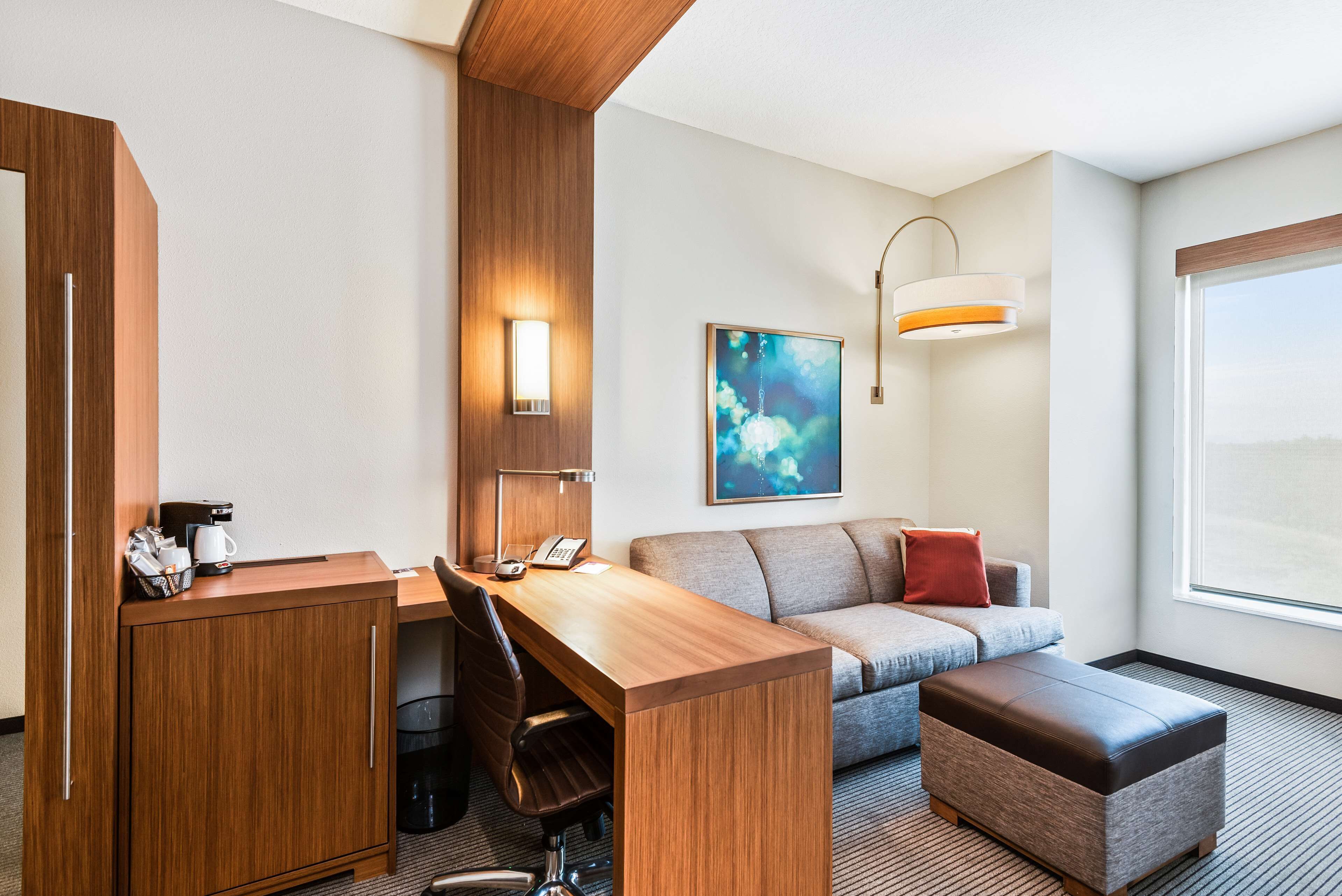 A hotel room with a wooden desk, a sofa, a cushioned bench, a coffee machine, a trash bin, and a painting on the wall.