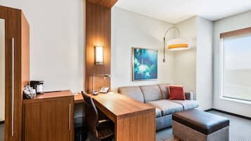 Une chambre d’hôtel avec un bureau en bois, un canapé, un banc rembourré, une machine à café, une poubelle et un tableau au mur.