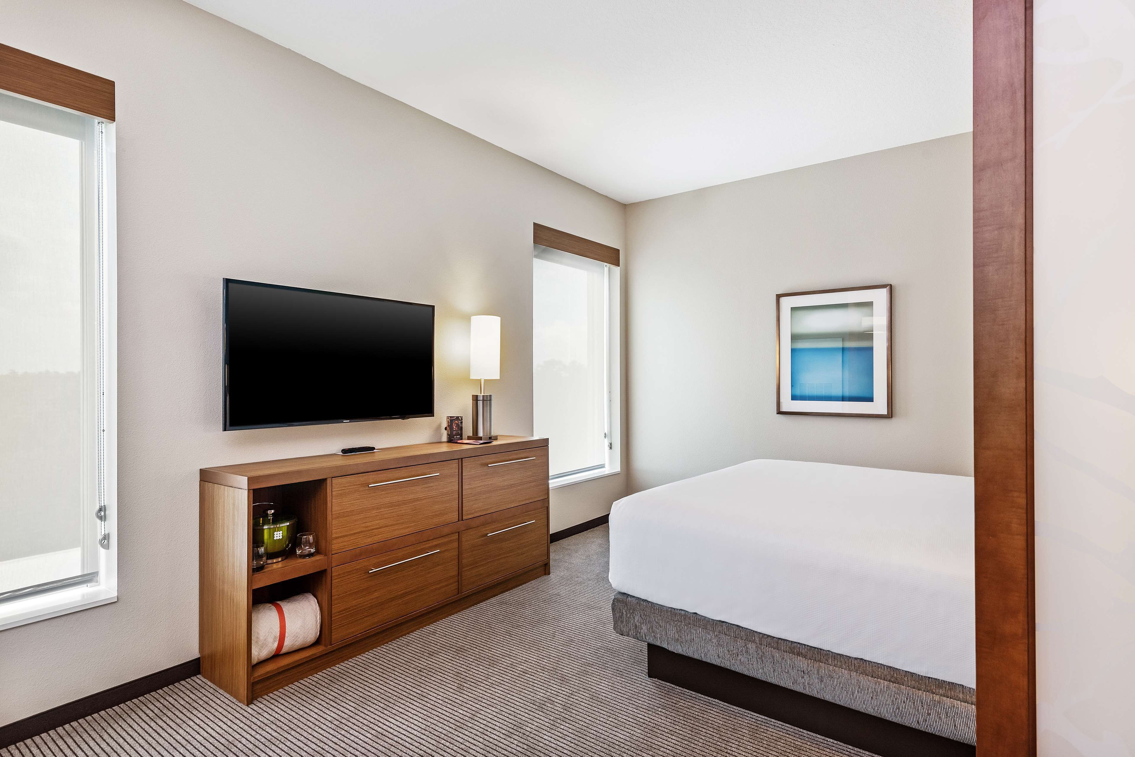 Une chambre d’hôtel avec un lit, un téléviseur fixé au mur, une commode en bois, une fenêtre et un tableau encadré au mur.