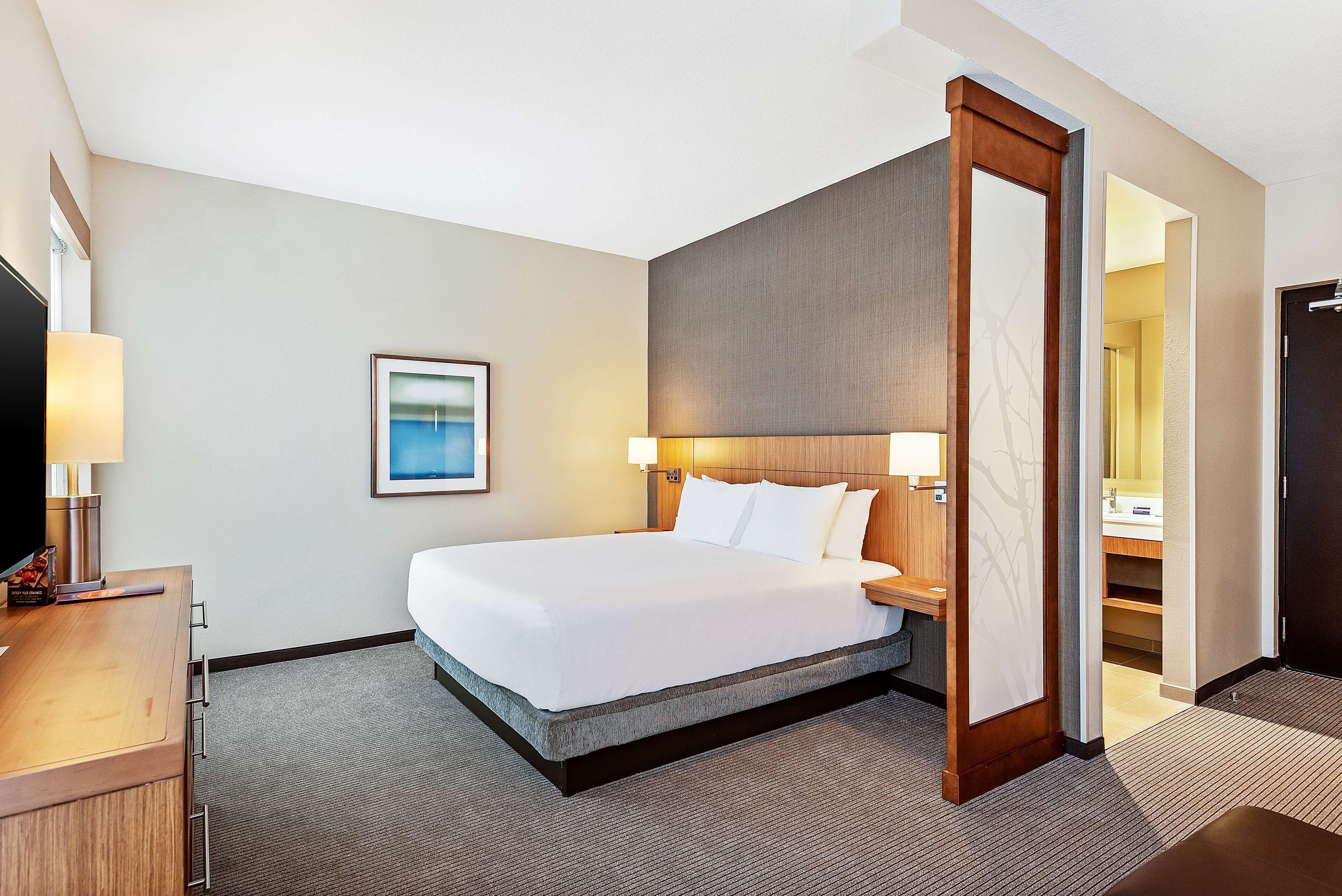 Une chambre d’hôtel avec un grand lit, des tables de chevet, un bureau avec une lampe, un tableau encadré au mur et une porte donnant sur une salle de bain.