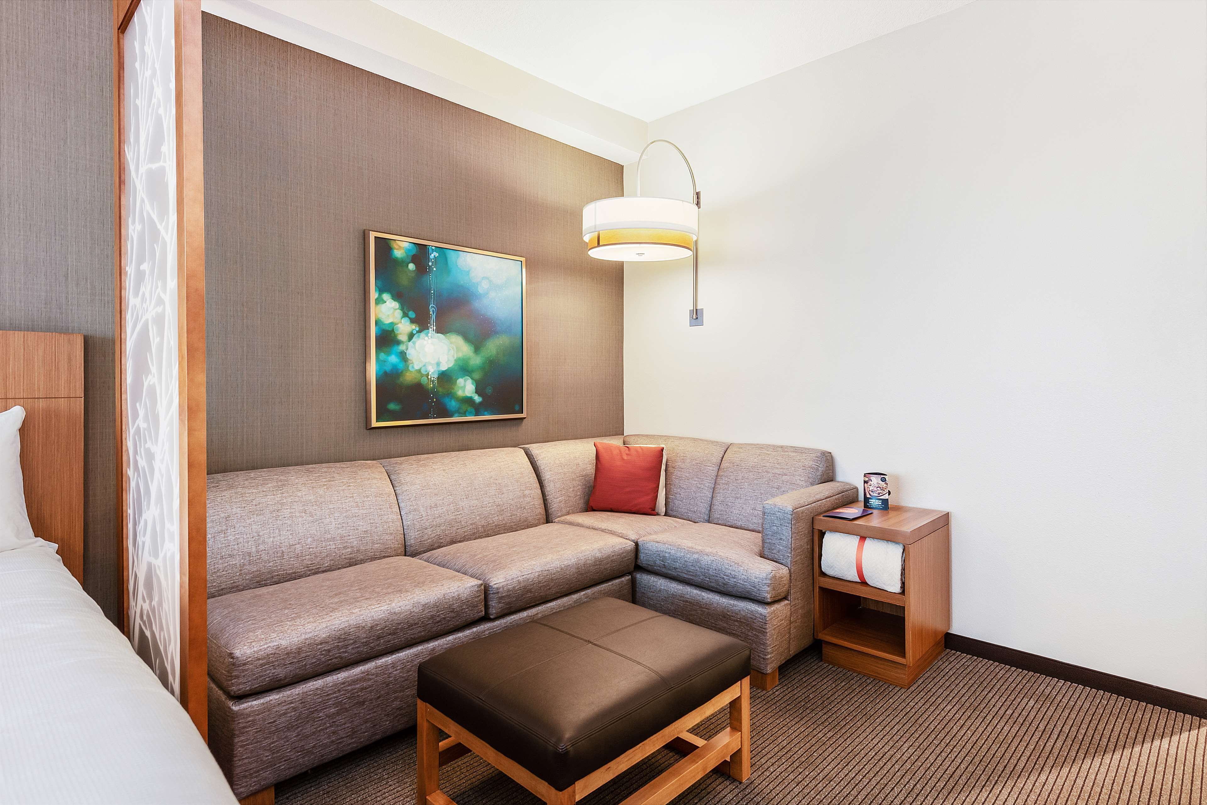 A hotel room with a sectional sofa, a wooden nightstand, a painting, and a bed.