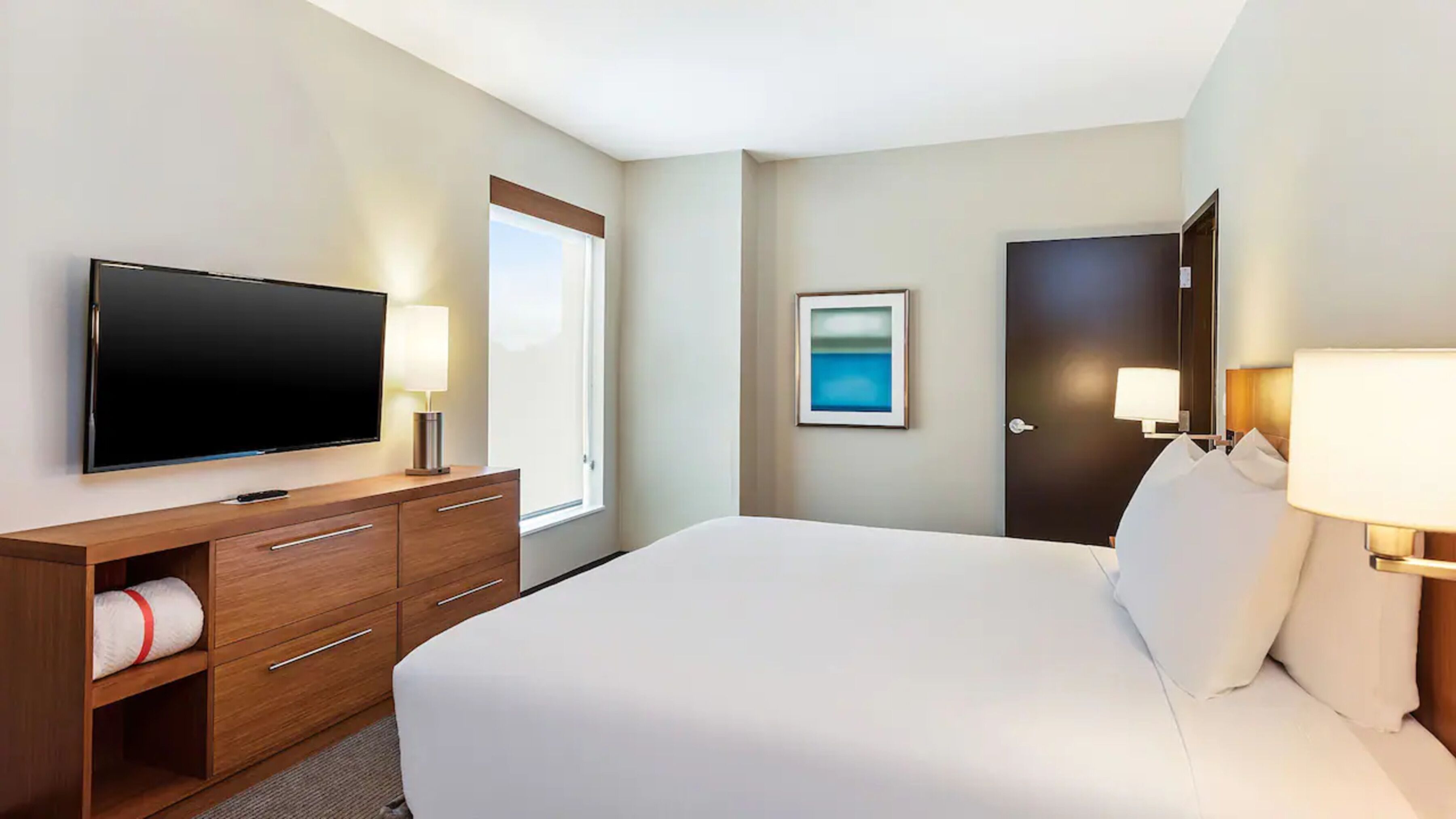 A hotel room with a large bed, a flat-screen TV, a wooden dresser, and a framed picture on the wall.