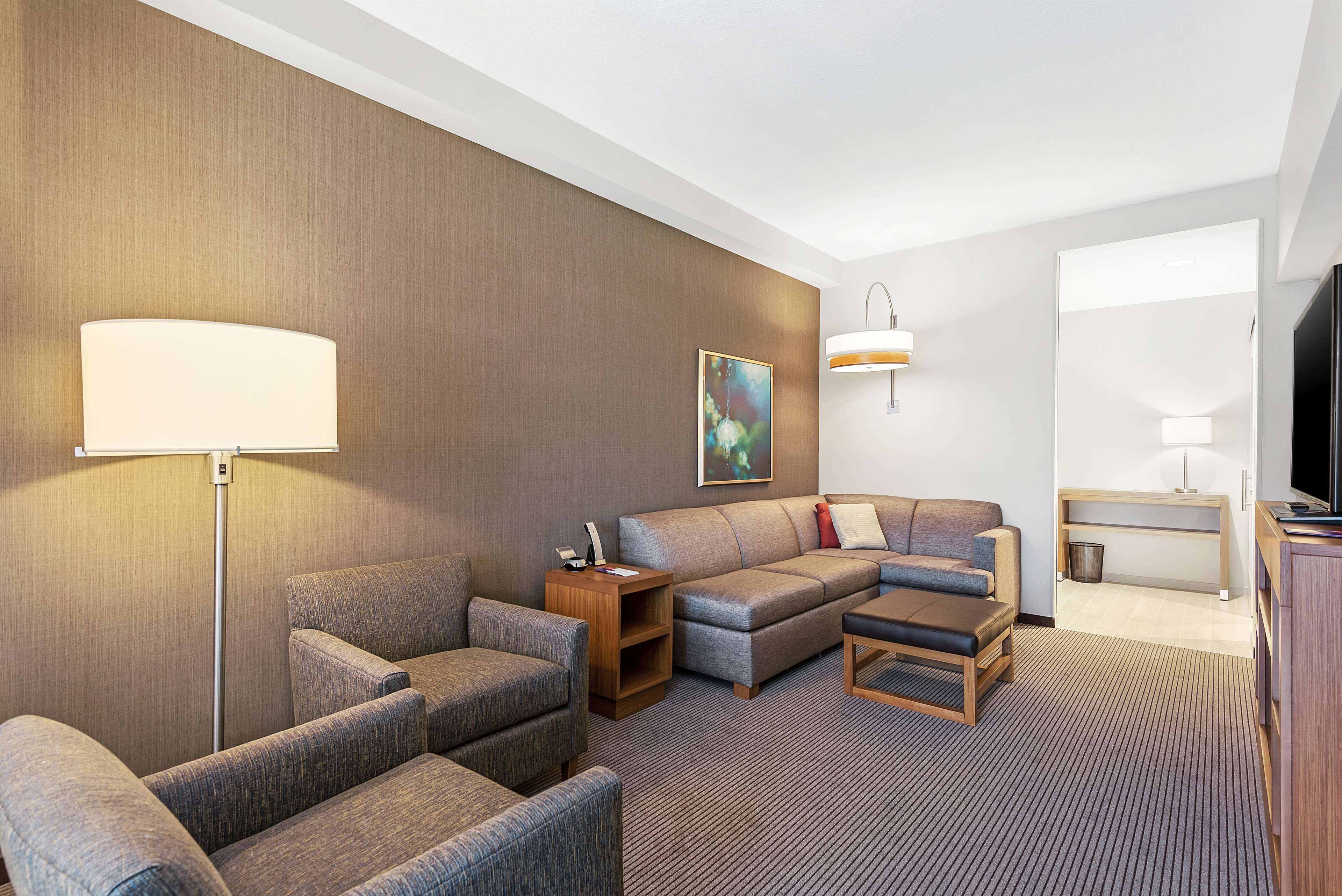 Habitación de hotel con sofá modular, sillones, una mesita, lámpara, un cuadro y televisor.