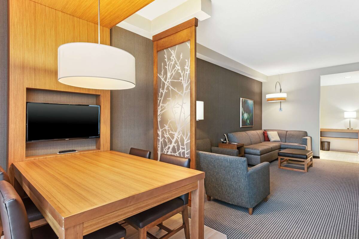 A hotel room with a wooden dining table, a flat-screen TV, a sofa, and a small coffee table.