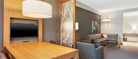 A hotel room with a wooden dining table, a flat-screen TV, a sofa, and a small coffee table.
