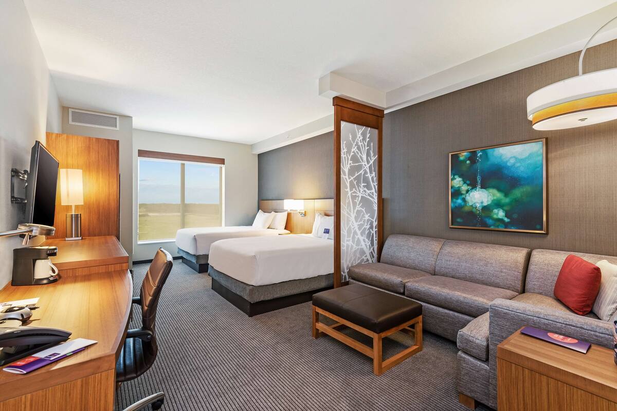 A hotel room with a bed, a sofa, a desk with a computer, a TV, and a painting on the wall.