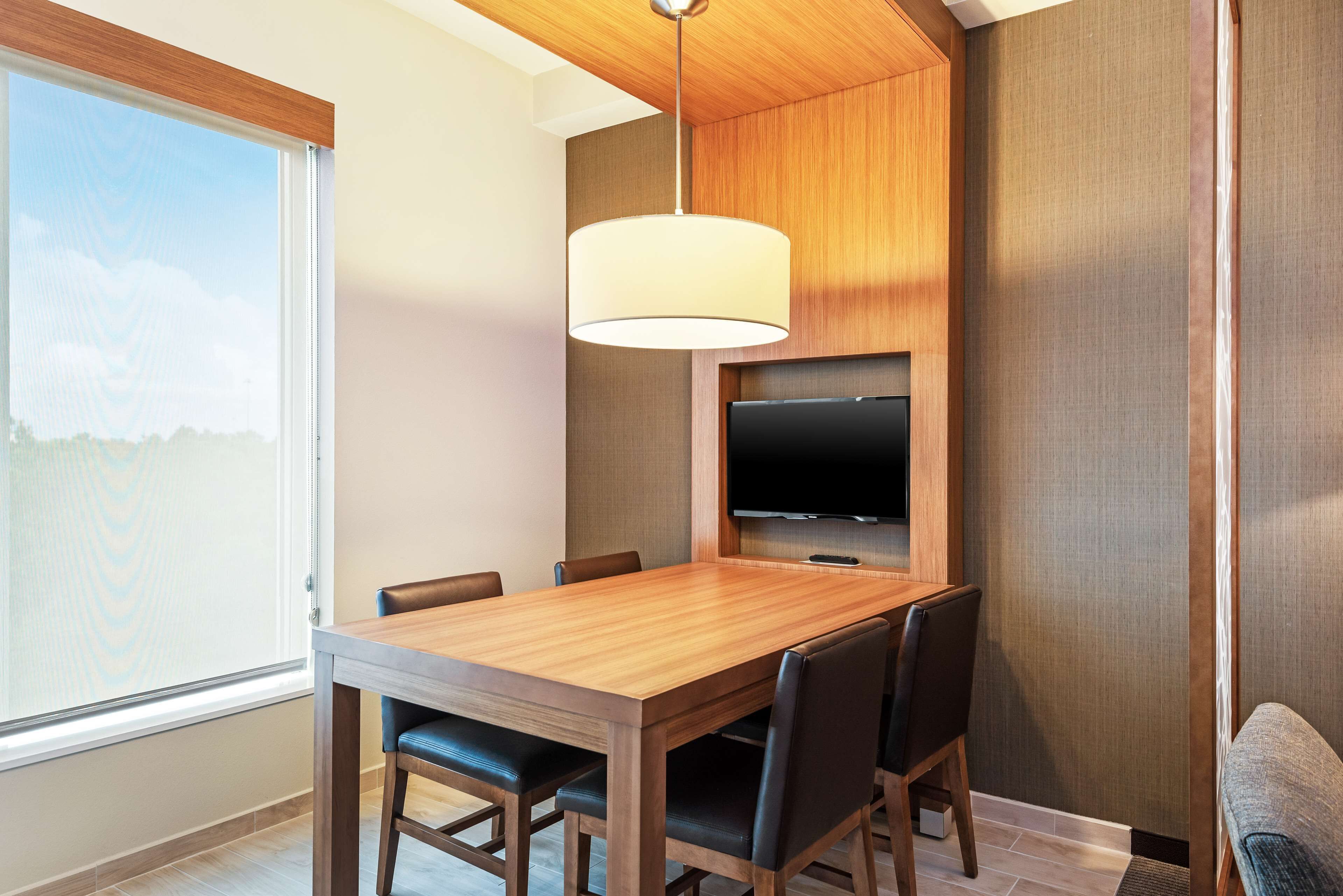 Un comedor moderno con una mesa de madera, seis sillas, un televisor montado en la pared y un ventanal que ofrece vistas al exterior.