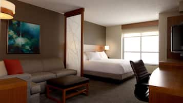 A hotel room with a bed, a sofa, a desk, a chair, a TV, and a painting on the wall.