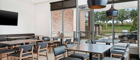 A spacious indoor dining area with multiple tables and chairs, large windows offering a view of the outdoors, and a flat-screen TV mounted on the wall.
