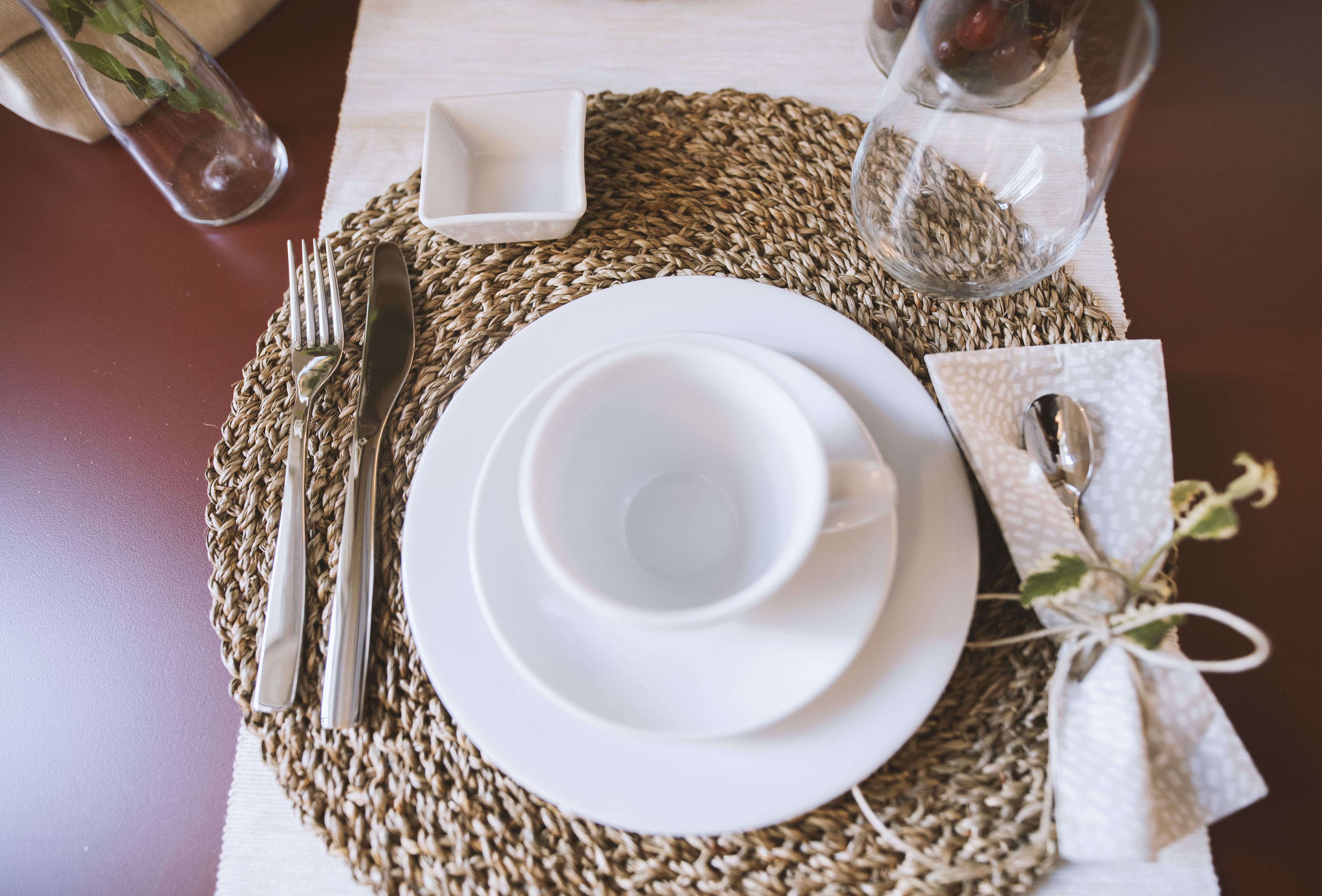 A neatly set dining table with white plates, cutlery, and a woven placemat.