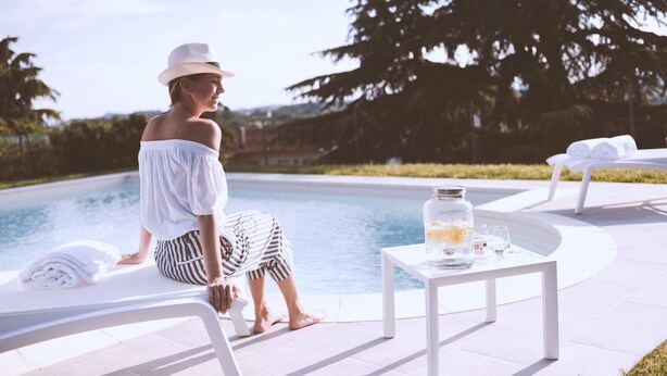 A person sitting by a poolside lounge chair with a table and a glass jar of drinks nearby. - Moonfrà (Antignano)