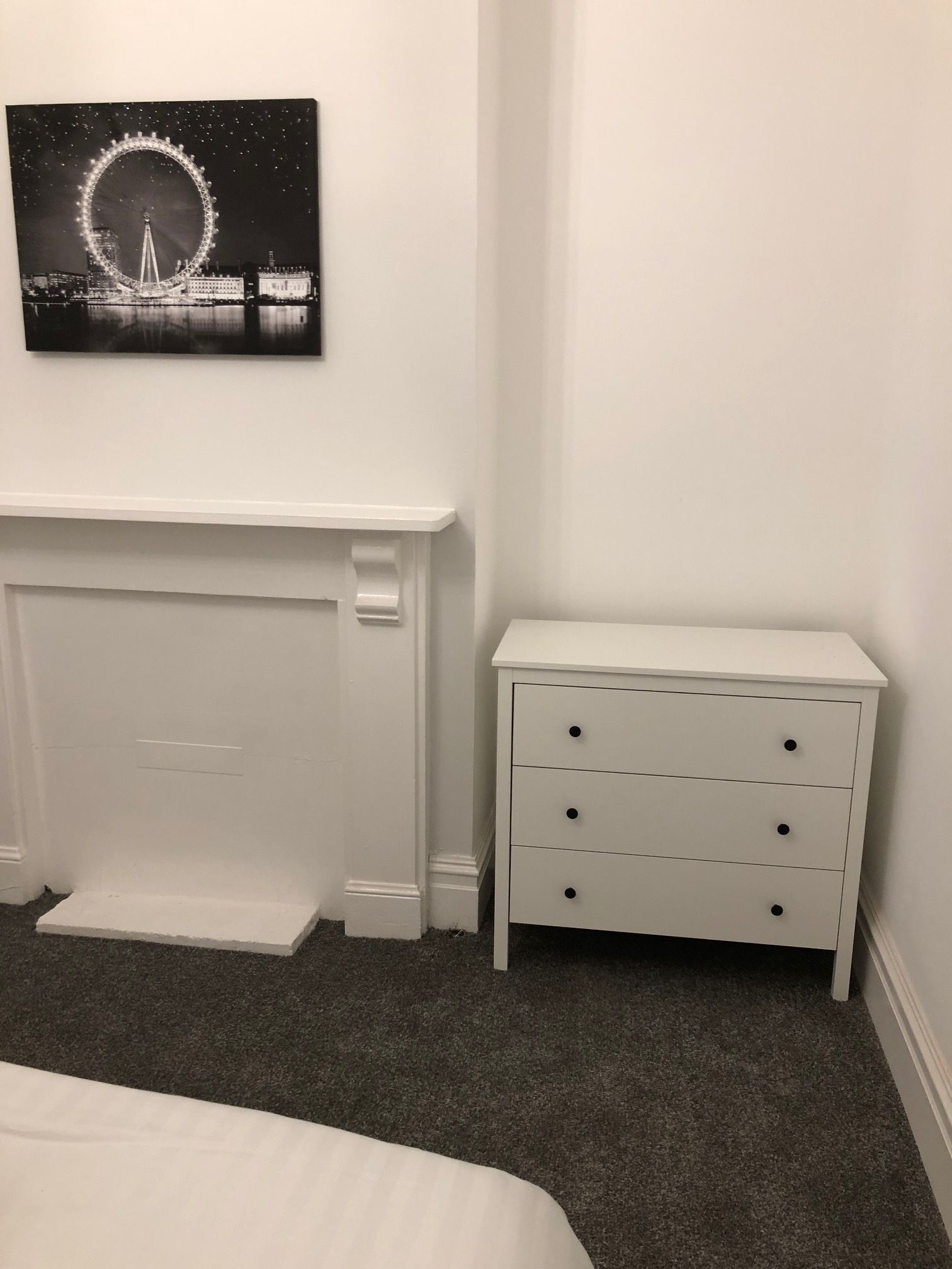 A bedroom with a white fireplace, a nightstand with drawers, and a wall-mounted picture of the London Eye.
