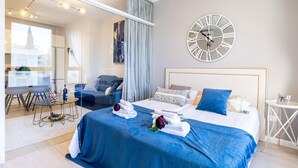 A modern hotel room with a large bed, a round wall clock, a dining area with a glass table, and a view of a building outside. - King David Court - Isrentals (Jerusalem)