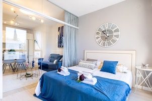 Habitación de hotel moderna con una cama grande, un reloj de pared redondo, una zona de comedor con una mesa de vidrio y vistas a un edificio.