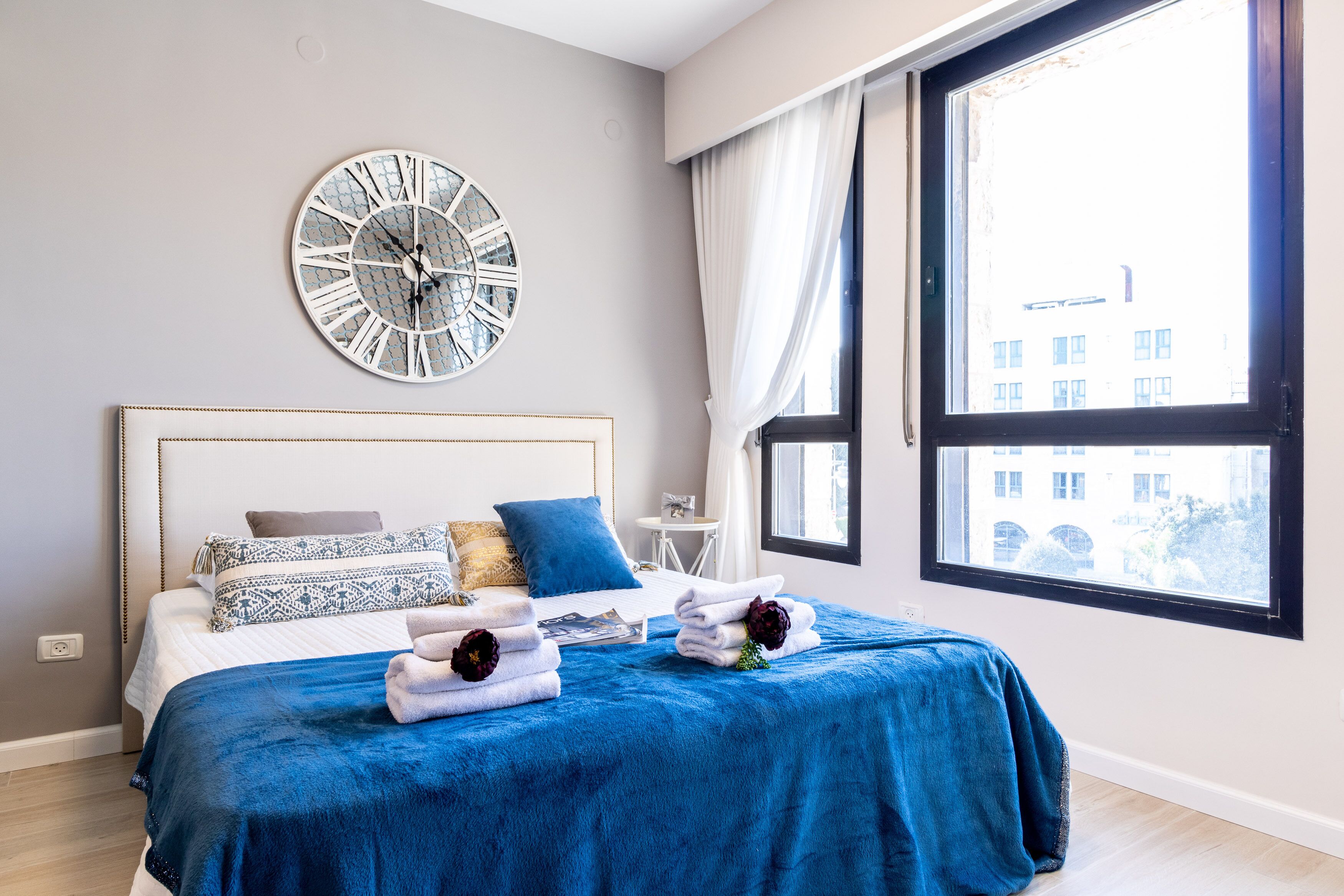 A modern bedroom with a large bed, a decorative clock, and a view of a building through a window.