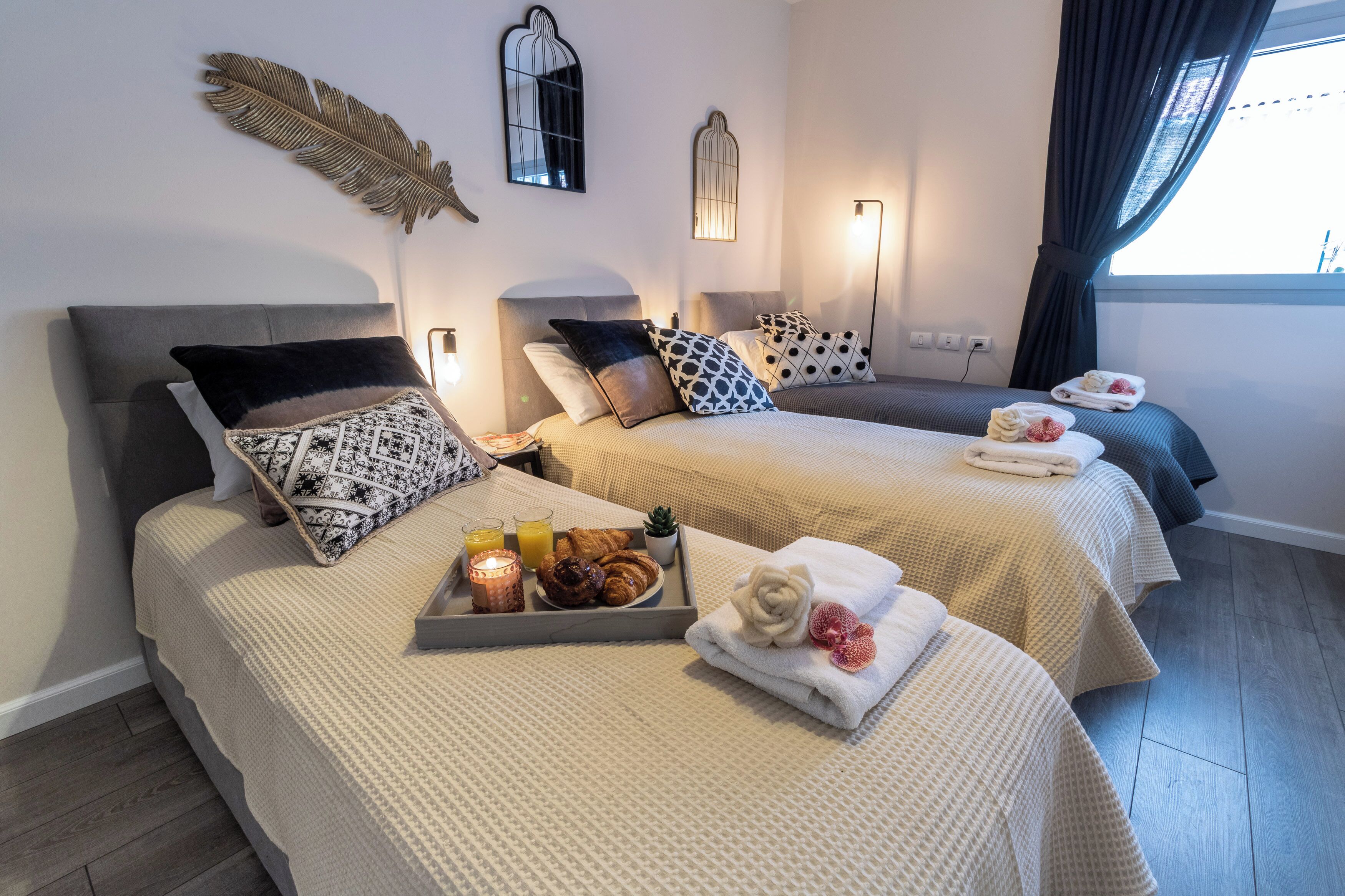 Habitación de hotel con dos camas, cada una con un cojín de diseño diferente, una bandeja con croissants y jugo, y toallas con motivos florales.