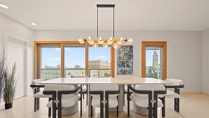 A modern dining area with a large table and six chairs, large windows with a city view, and a hanging light fixture. - King David Court - Isrentals (Jerusalem)