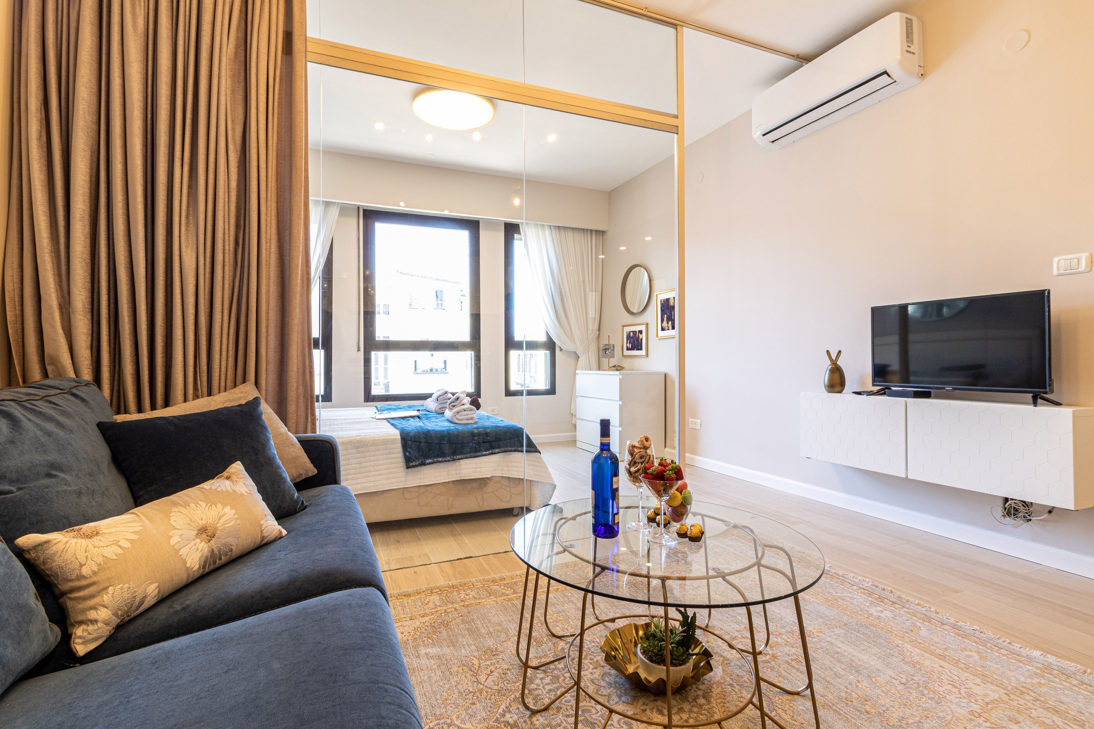 Una sala de estar moderna con un sofá, una mesa de centro de vidrio con objetos decorativos, un televisor sobre un mueble blanco y un ventanal con cortinas.