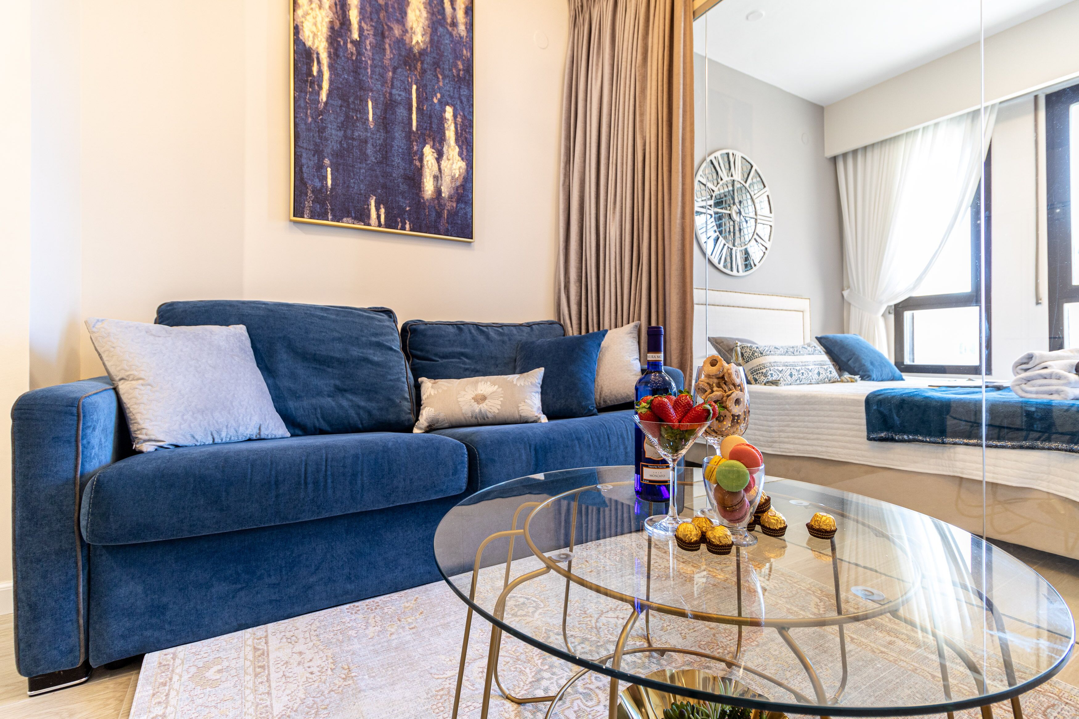 Een moderne woonkamer met een blauwe bank, een glazen bijzettafeltje met fruit en gebak, een groot abstract schilderij en een klok aan de muur.