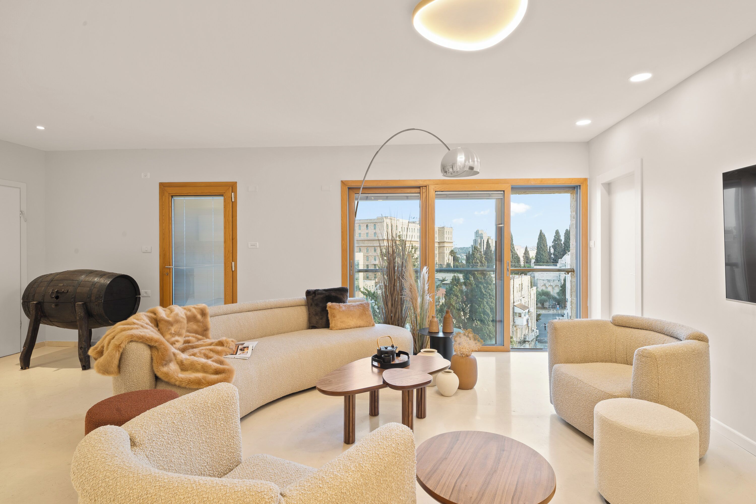 A modern living room with a sofa, armchairs, and a coffee table. There is a large window with a view of buildings and trees.