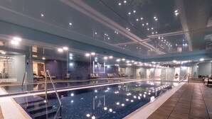 An indoor swimming pool with clear water, surrounded by a wooden deck and glass walls. - King David Court - Isrentals (Jerusalem)