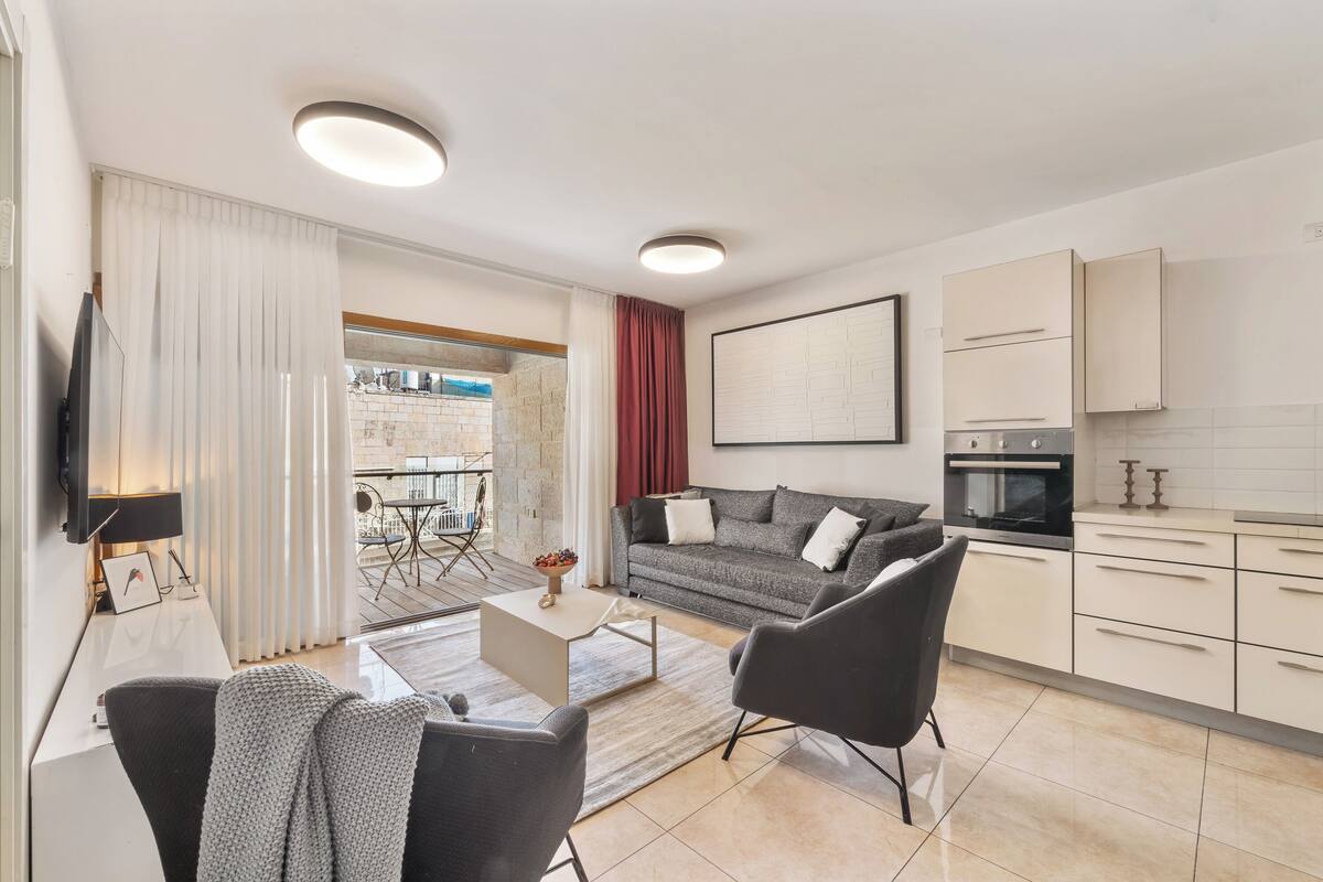 Ein modernes Wohnzimmer mit einer Couch, Sesseln, einem Couchtisch und einem Fernseher. Es gibt eine Küchenzeile mit Schränken und einem Kühlschrank.