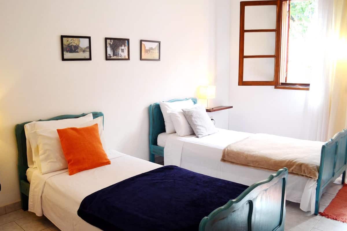 A room with two beds, each with a different colored headboard, a window with a wooden frame, and three framed pictures on the wall.