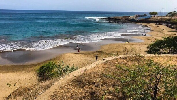 A beach with clear blue water, sandy shore, and some vegetation. - Syds Guesthouse I (Praia)