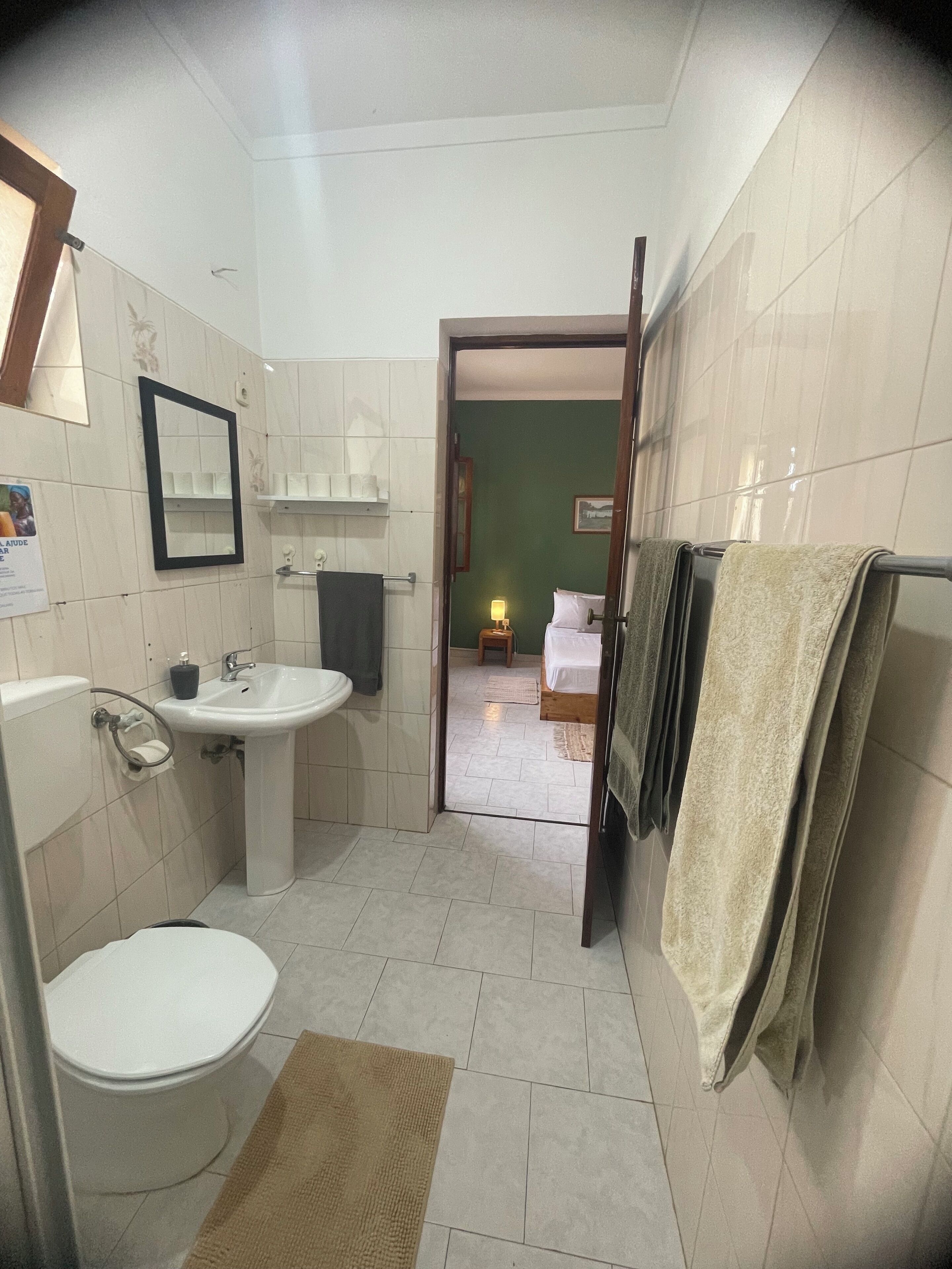 A bathroom with a toilet, sink, and mirror. There is a towel rack with towels and a small stool in the adjacent room.